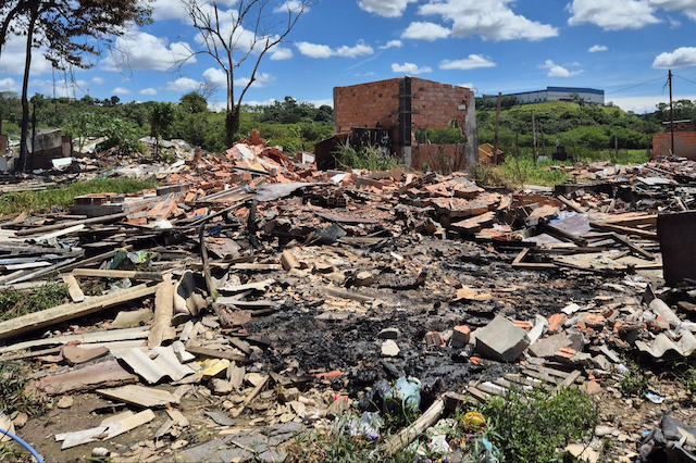 Um terreno a ceu aberto repleto de entulho, escombros e restos de contrução com uma construção ainda em fase de demolição mais ao fundo com uma árvore seca e algumas vegetações rasteiras mais ao fundo.